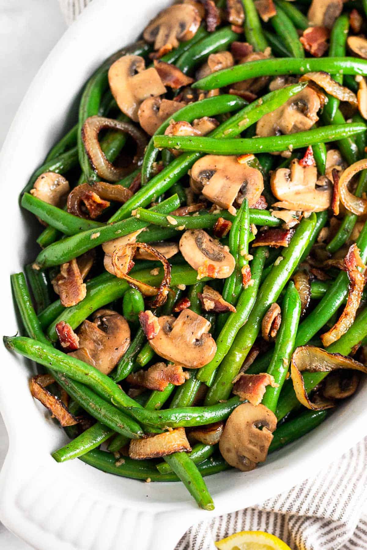 Green beans and mushrooms with bacon and fried onions in a white casserole dish. The dish is sitting on a striped towel with a lemon wedge next to it.