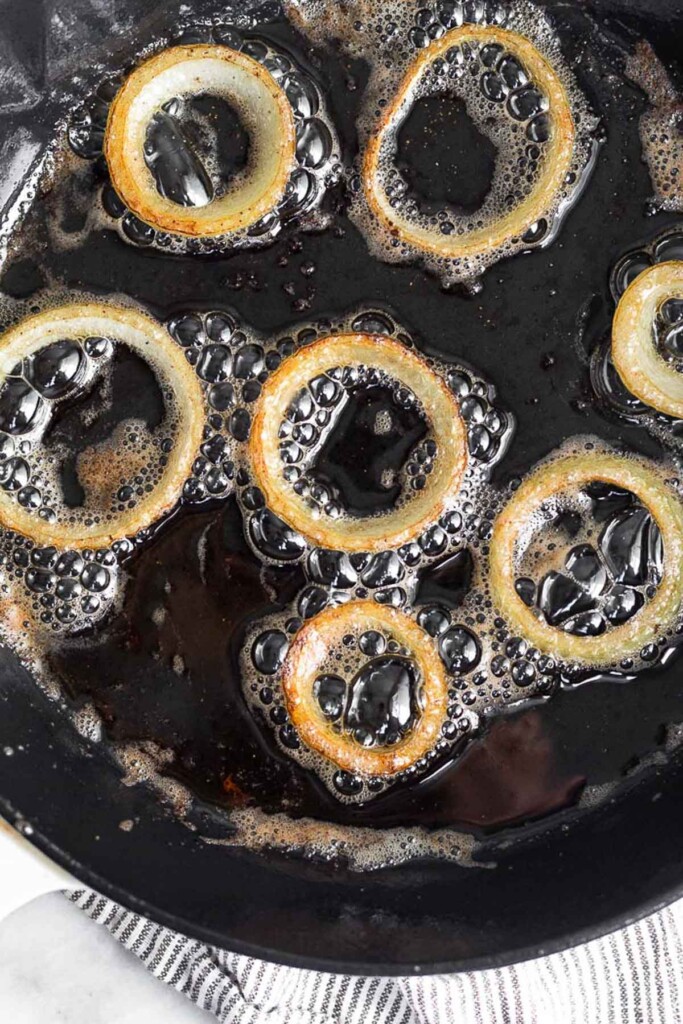 Overhead shot of cast iron skillet with rings of onions frying in oil in it.