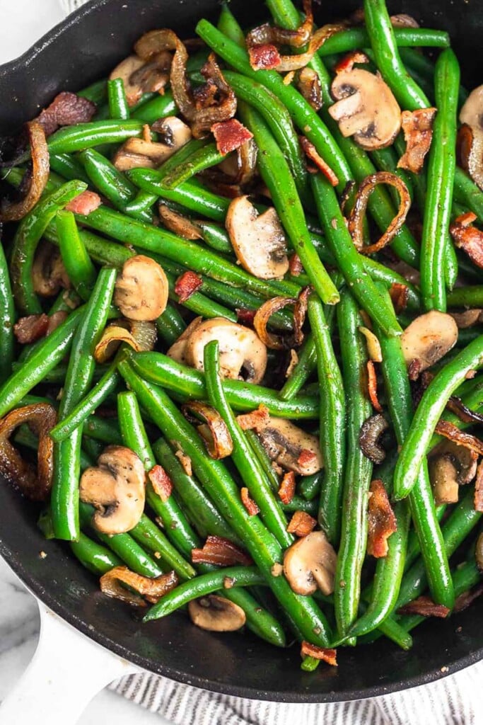 Sautéed green beans with mushrooms and onions topped with bacon pieces in a large cast iron skillet.