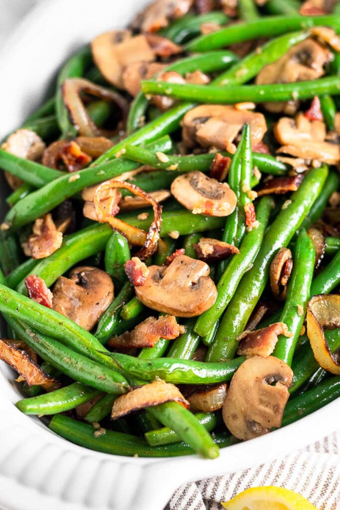 Close up shot of sautéed mushrooms and green beans with onions and bacon in a white casserole dish.