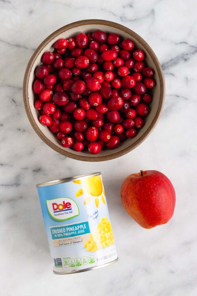 White marble counter with a bowl of fresh cranberries, a whole red apple, and a canned of crushed pineapple.