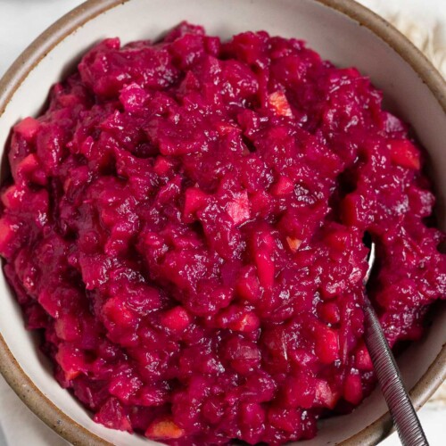White bowl of homemade cranberry sauce with a spoon in it. Behind it is a bowl of fresh cranberries.