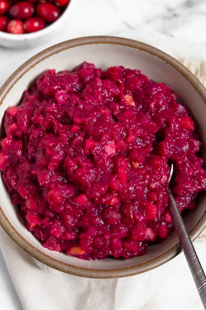 White bowl of homemade cranberry sauce with a spoon in it. Behind it is a bowl of fresh cranberries.