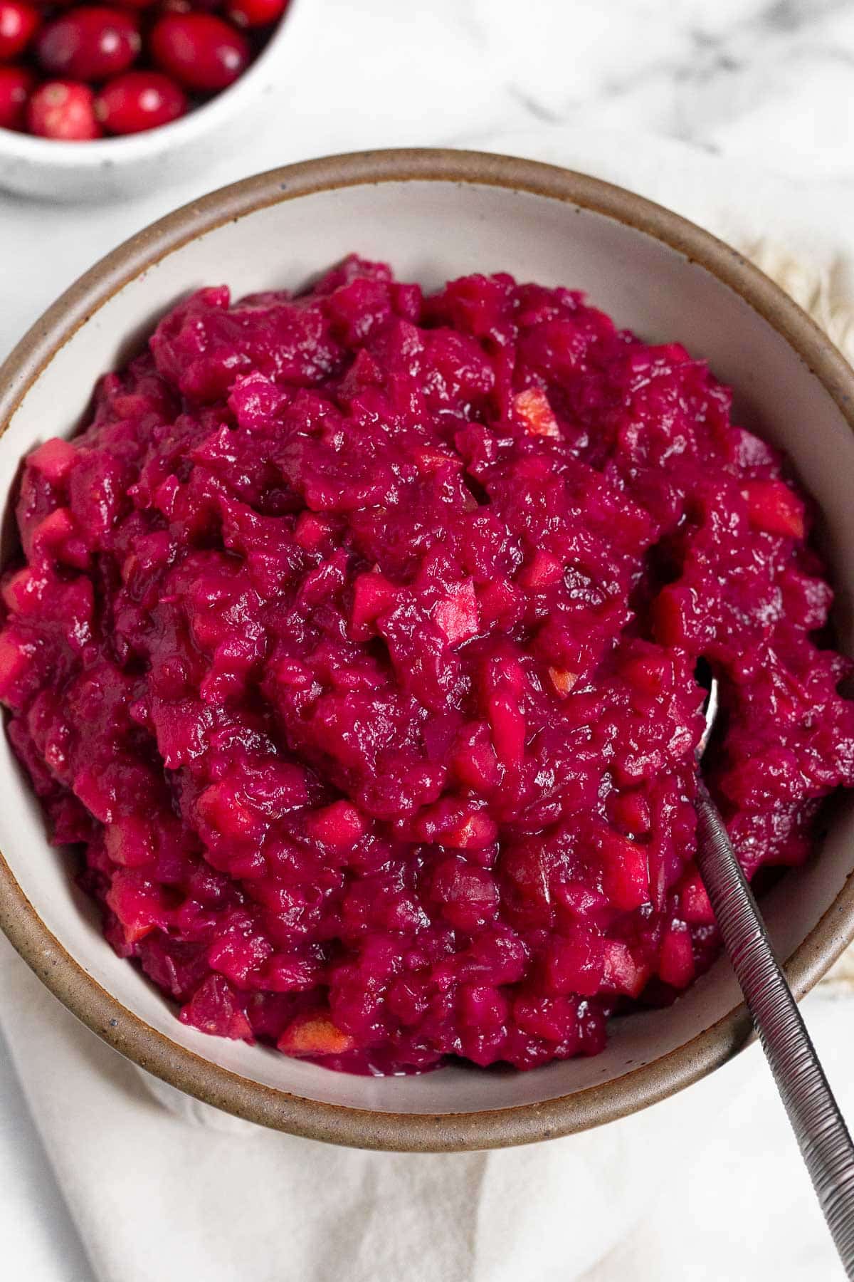 White bowl of homemade cranberry sauce with a spoon in it. Behind it is a bowl of fresh cranberries.