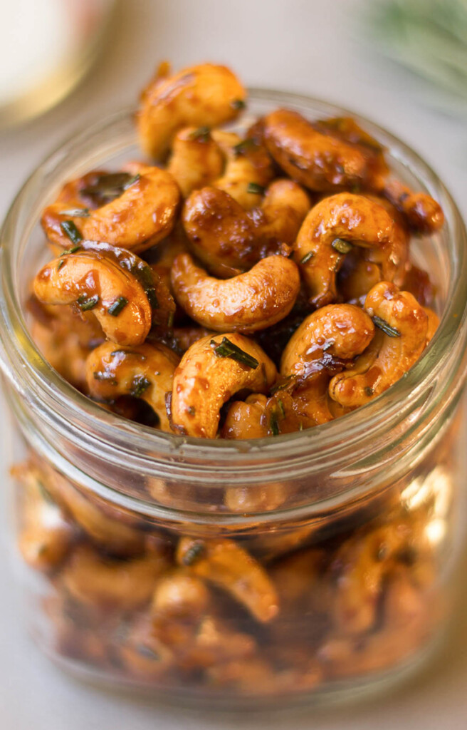 Jar filled with rosemary cashews. In the background is a sprig of fresh rosemary.