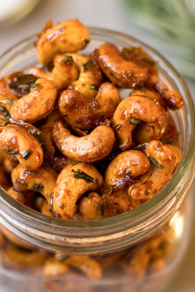 Close up overhead shot of honey roasted cashews in a glass jar. In the background is a sprig of fresh rosemary.