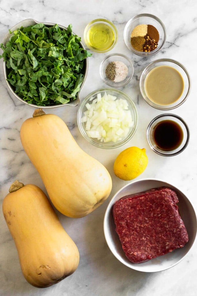 White marble counter with a small bowl of oil, a bowl of spices, a bowl of tahini, a bowl of salt and pepper, a bowl of coconut aminos, a bowl of diced onion, a whole lemon, a bowl of raw ground beef, two butternut squashes, and a bowl of chopped kale.
