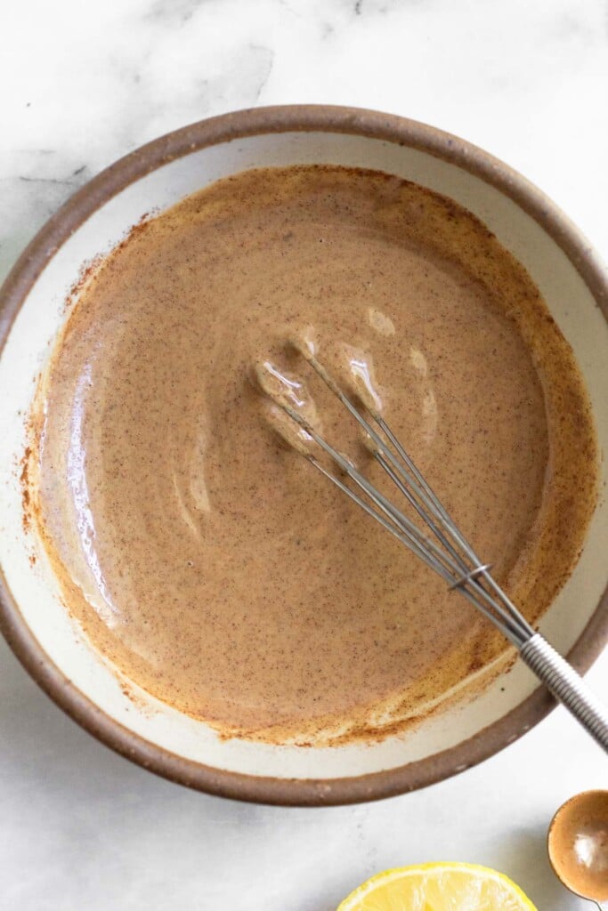 A bowl of tahini sauce with a small whisk in it. Next to the bowl is half a lemon and teaspoon.