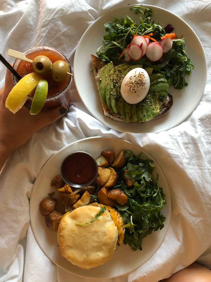 Room service of two plates of breakfast on top of a bed with a bloody mary next to one of them.