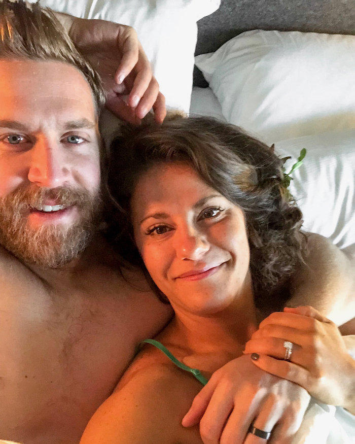 Husband and wife in bed the day after they got married. The wife is laying on husband's shoulder and they are both smiling.