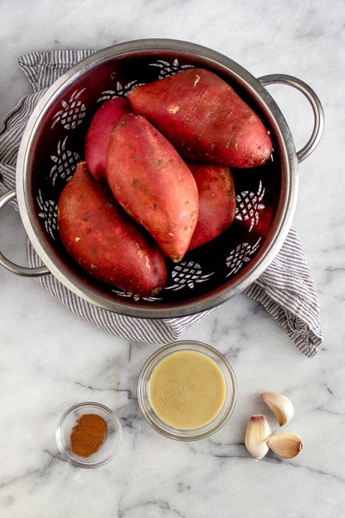 White marble counter with a colander of sweet potatoes, a bowl of tahini, three cloves of garlic, and a bowl of cinnamon.