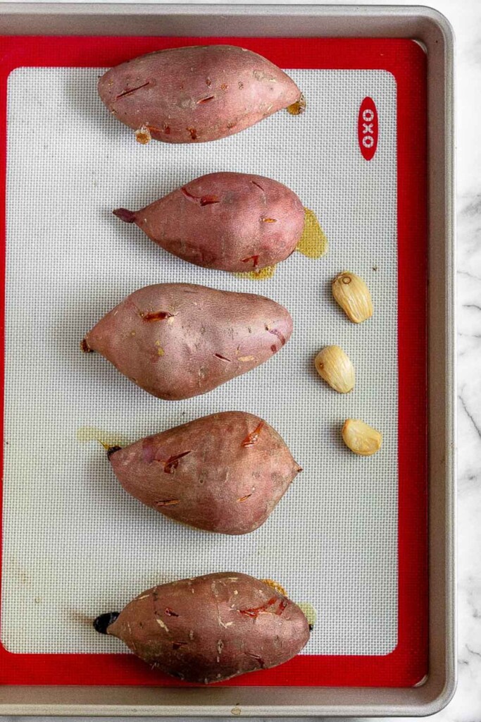 Baking sheet with 5 roasted sweet potatoes and 3 roasted cloves of garlic on it.