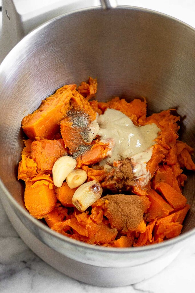 Large metal bowl filled with roughly chopped roasted sweet potatoes, tahini, roasted garlic, cinnamon, and salt and pepper.