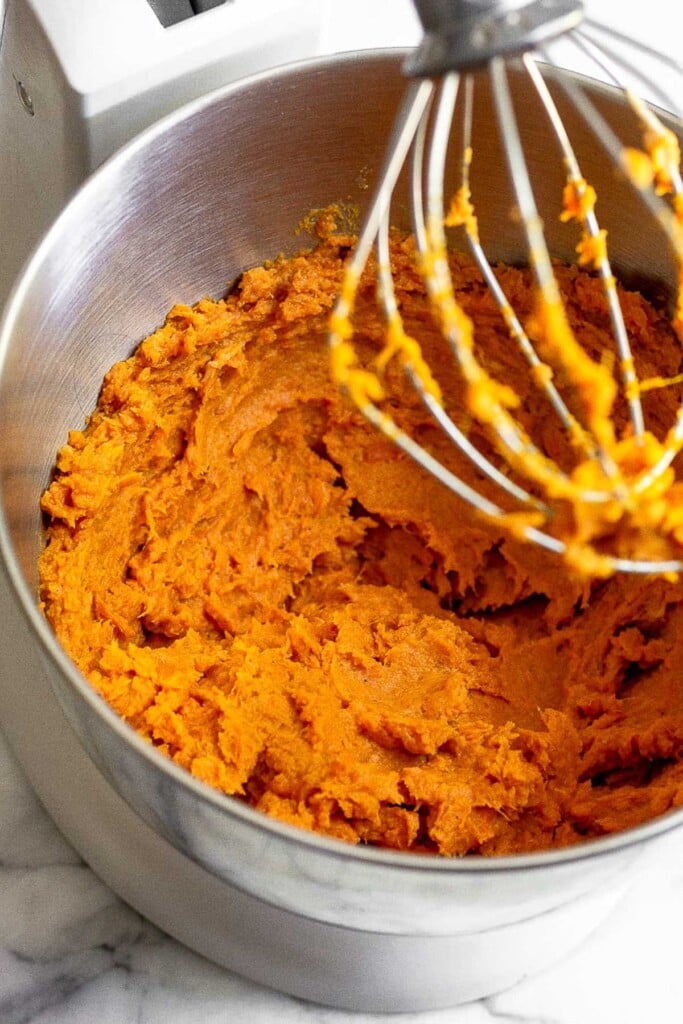 A stand mixer bowl filled with mashed sweet potatoes. Overtop is the whisk attachment.