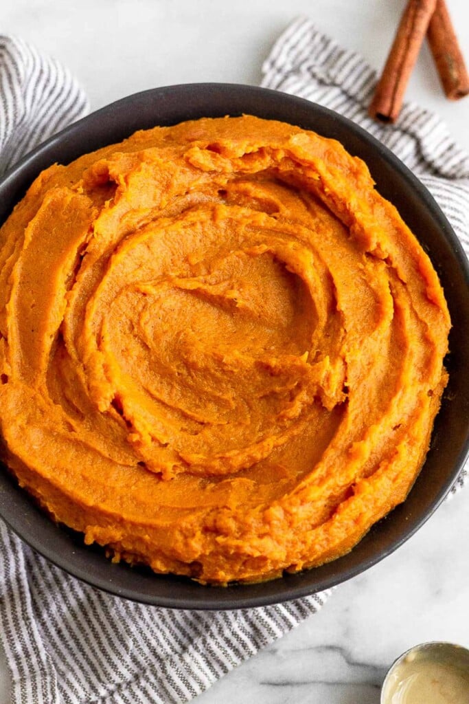 Mashed sweet potatoes in a black bowl. The bowl is sitting on a striped towel and next to it is a spoonful of tahini and cinnamon sticks.