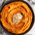 Bowl filled with whipped sweet potatoes topped with tahini and cinnamon. The bowl is sitting on a striped towel and next to it is a spoonful of tahini and cinnamon sticks.