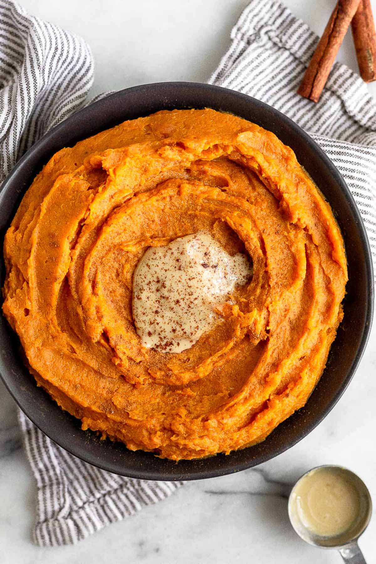 Bowl filled with whipped sweet potatoes topped with tahini and cinnamon. The bowl is sitting on a striped towel and next to it is a spoonful of tahini and cinnamon sticks.