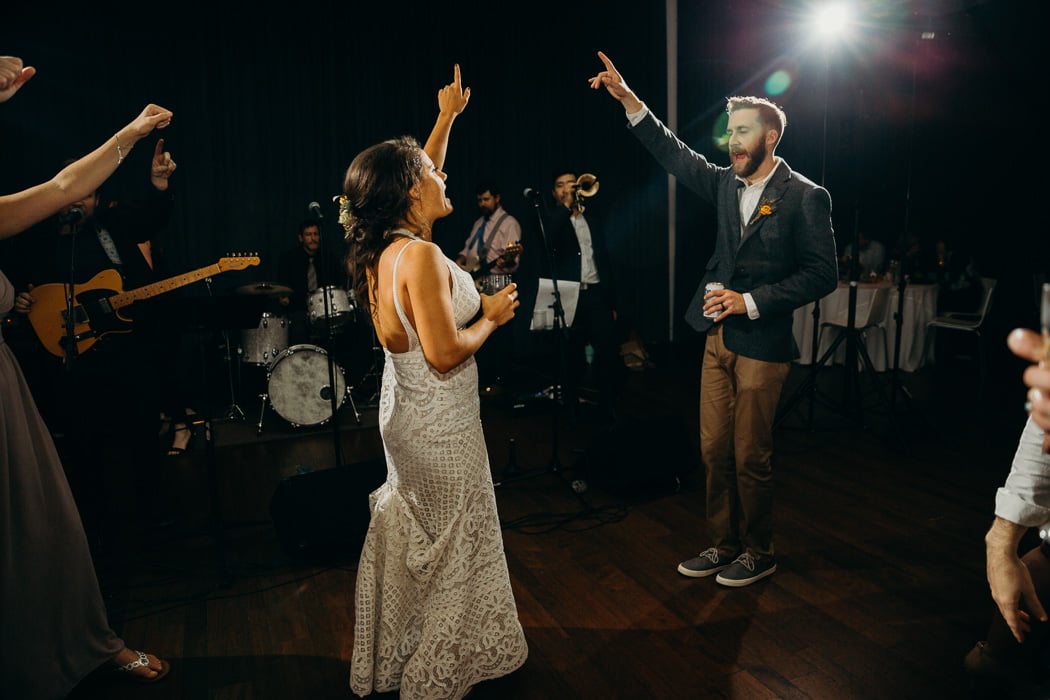 Both the bride and groom dancing on the dance floor and one hand up in the air and their finger pointed dancing and singing. A drink is in their other hand.
