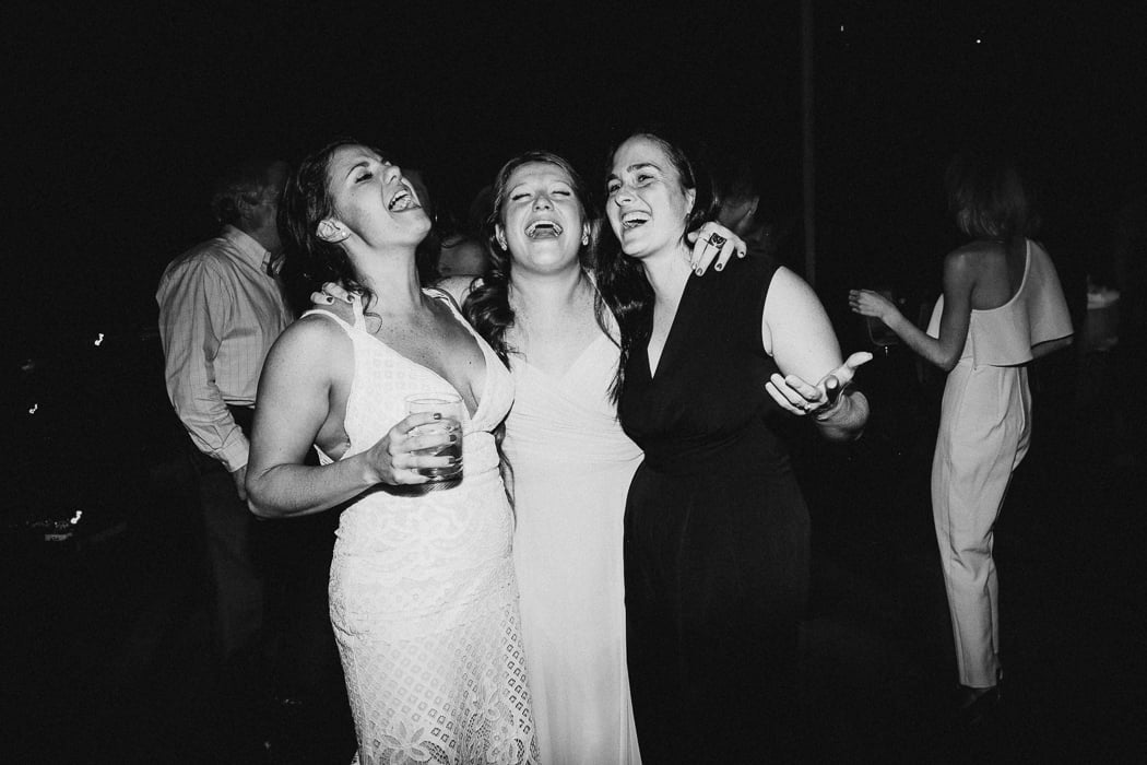 Three girls embracing at a wedding singing, one of them being the bride.