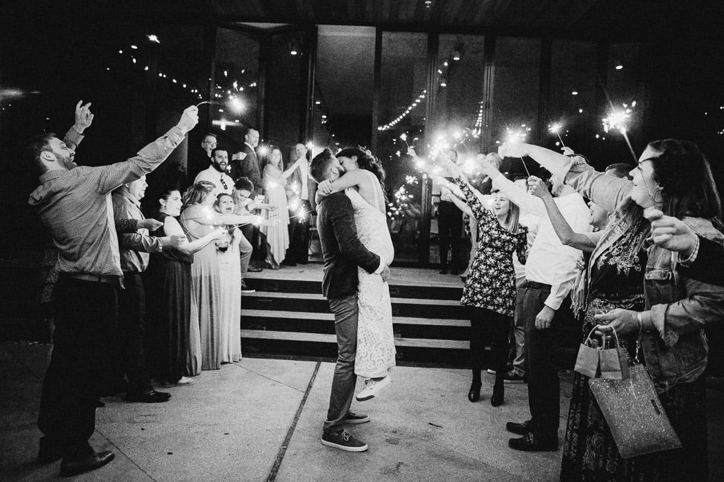 Bride and groom in the middle of two lines of people with sparklers. The groom is picking up the bride and they are kissing.