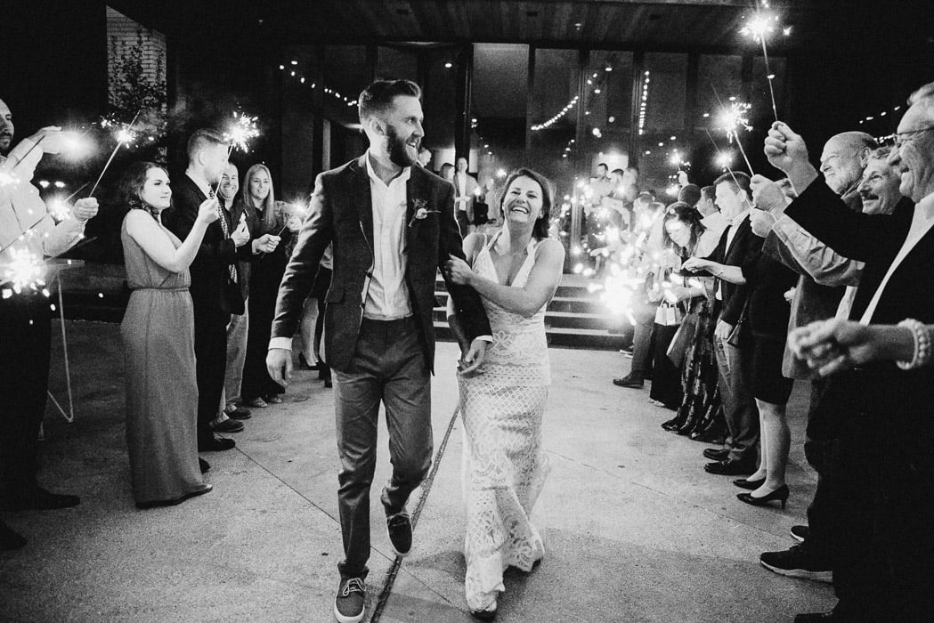 Bride and groom doing their exit of the ceremony through a line of sparklers. The bride has a huge smile on her face and the groom is looking over at the crowd.