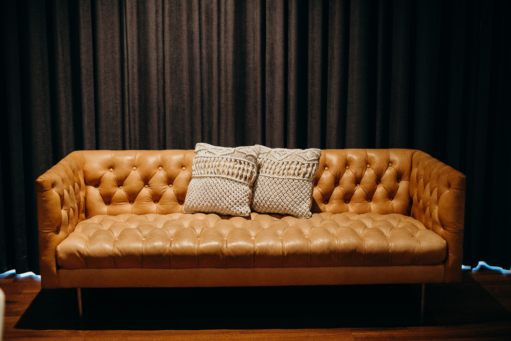 Brown leather couch with two pillows in front of a black curtain.