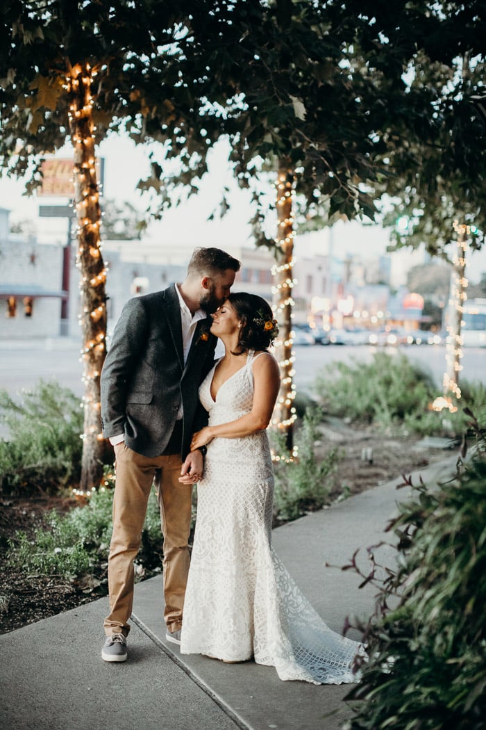 Bridge and groom standing on a sidewalk with greenery and lights around them. The groom is kissing the bride on her forehead.