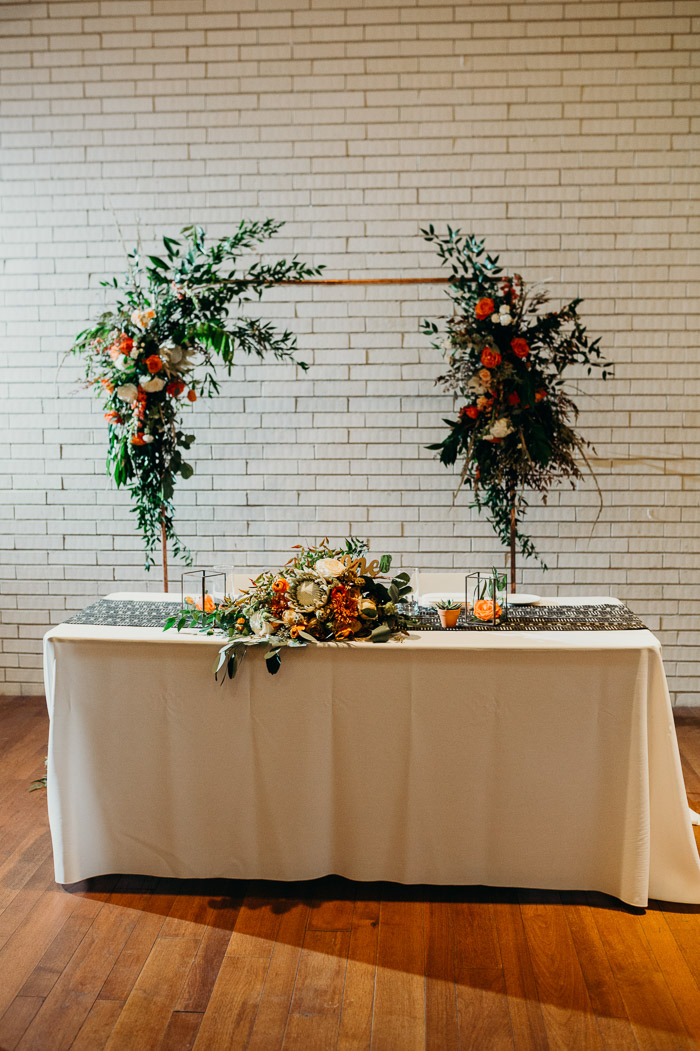 Bride and groom table at a wedding with the bouquet on it and an arch behind it with flowers. Behind it is a white brick wall.