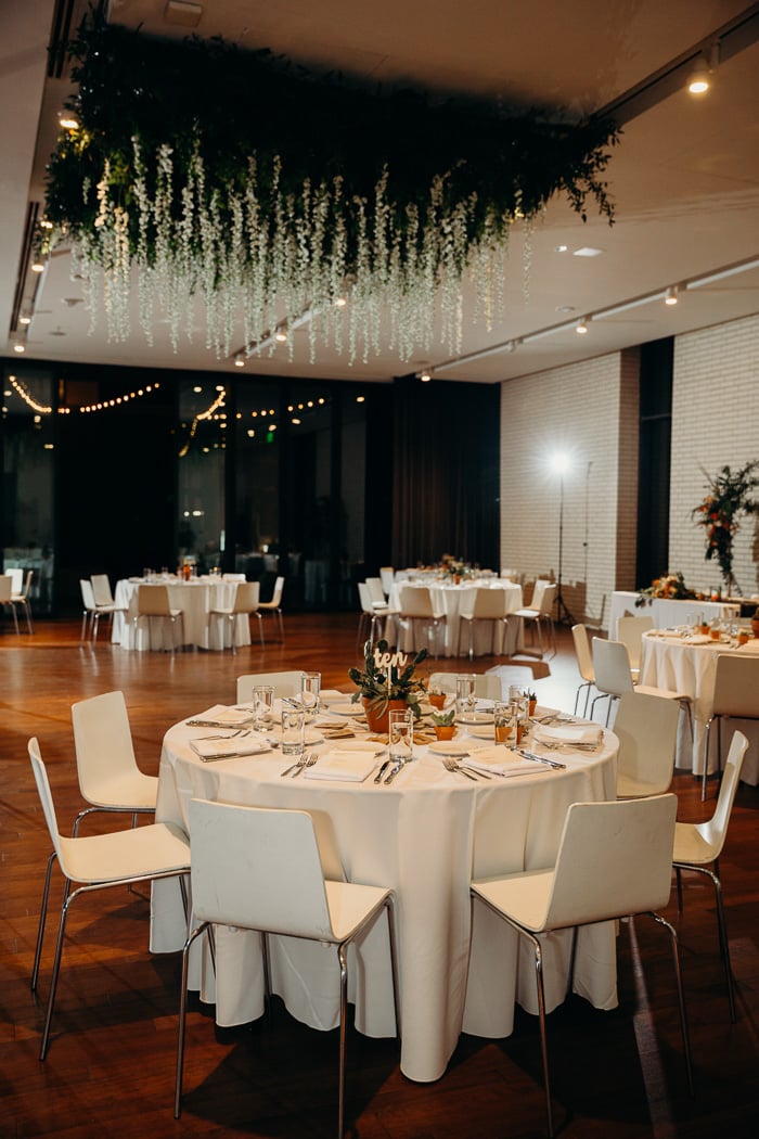 Tables and chairs set up for a wedding reception at South Congress Hotel. There is a large hanging from the ceiling with greens and flowers.