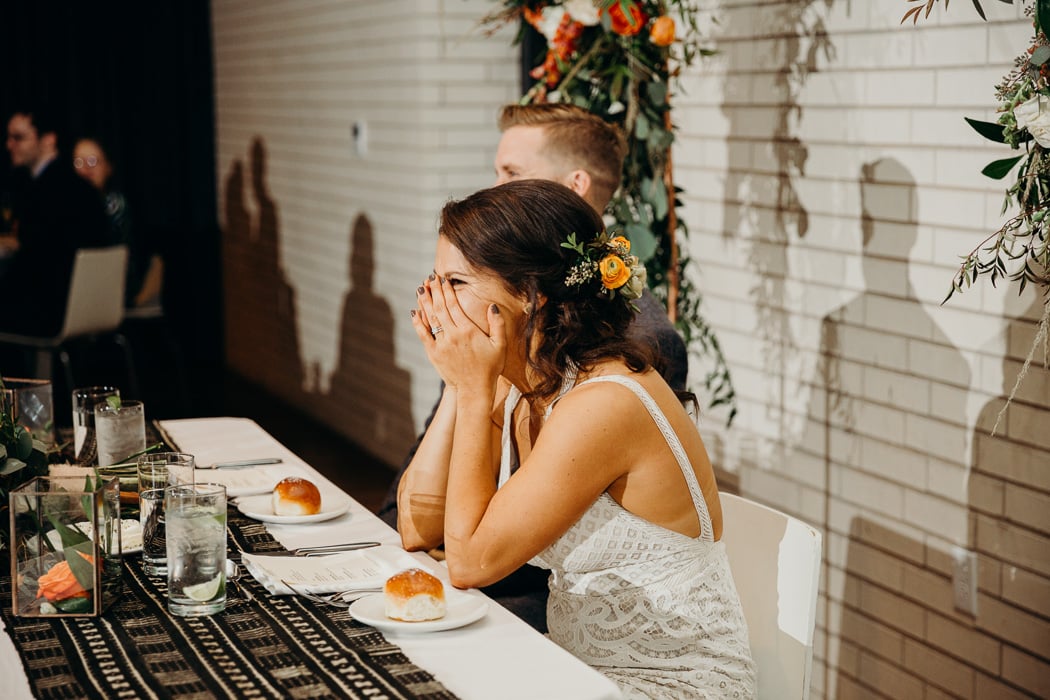 Bride and groom at the couple's table while someone is giving a speech. The bride's hands are over her mouth as she is laughing.