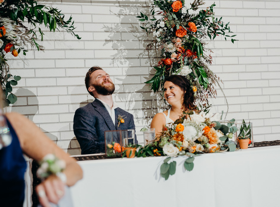 Bride and groom seating at their couples table on their wedding day. The groom has his head back laughing and the bride is looking at him smiling.