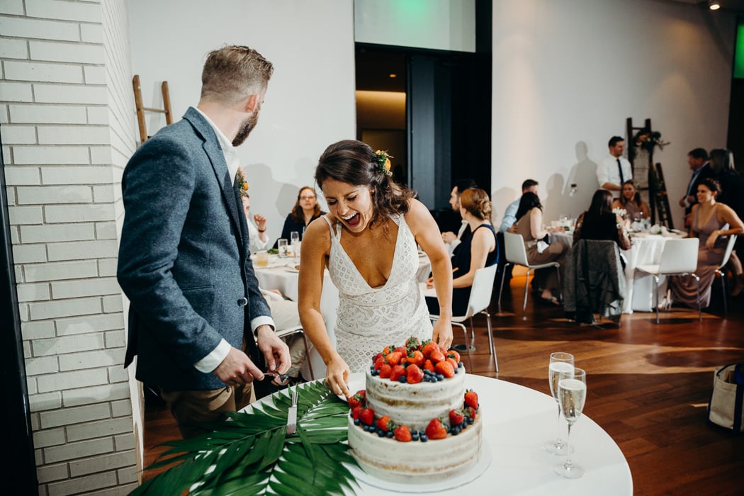 Bride and groom cutting their wedding cake. The groom is looking behind him and then bride is laughing. The cake is decorated with strawberries and blueberries.