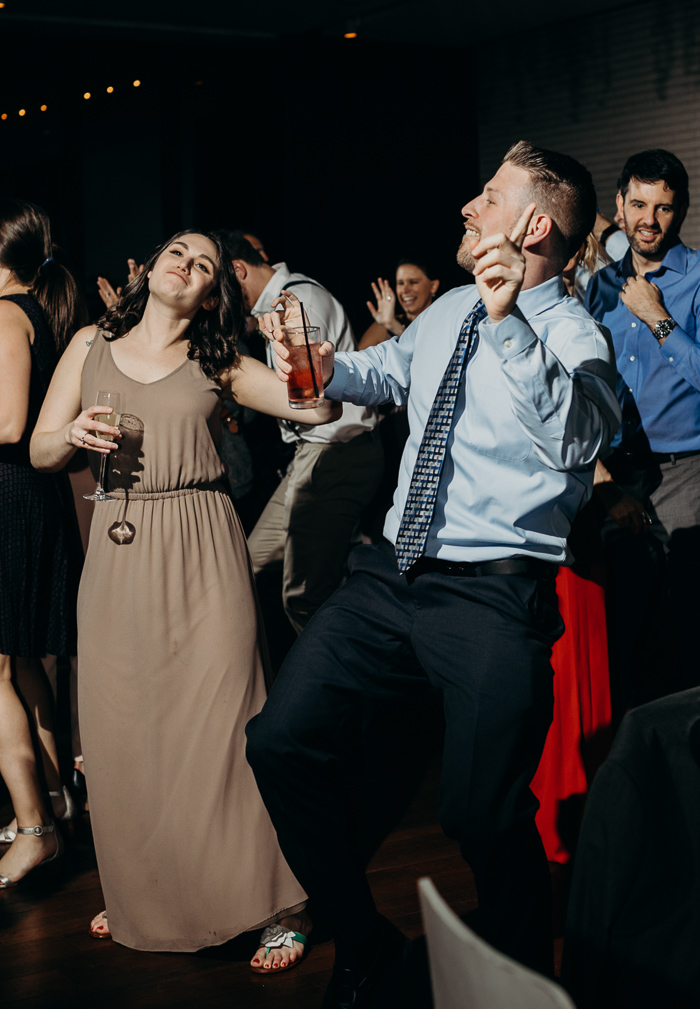 Guy and a girl with their one hand up and they other one holding a drink dancing at a wedding