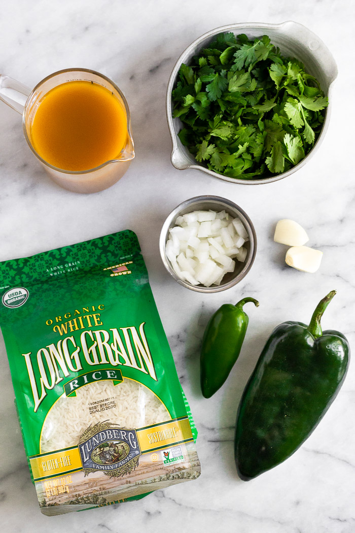 White counter with a bag of long grain white rice, cup of broth, bowl of fresh herbs, bowl of chopped onion, garlic cloves, and a poblano and jalapeno.