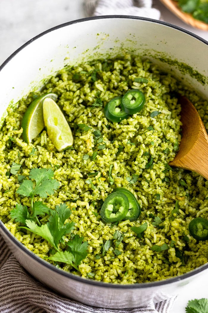 Close up of a large stock pot filled with arroz verde topped with jalapeños, limes, and cilantro. A wooden spoon is sticking out of it.