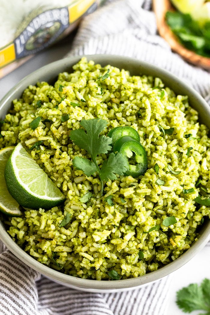 Grey bowl filled with green rice topped with lime wedges, cilantro, and sliced jalapenos. It is sitting on a stripped towel and behind it is a bag of white rice and a plate of herbs and limes.