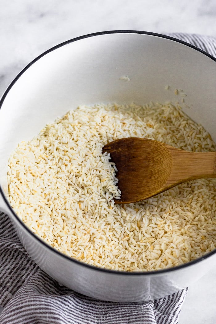Large white dutch oven filled with toasted rice with a wooden spoon in it.