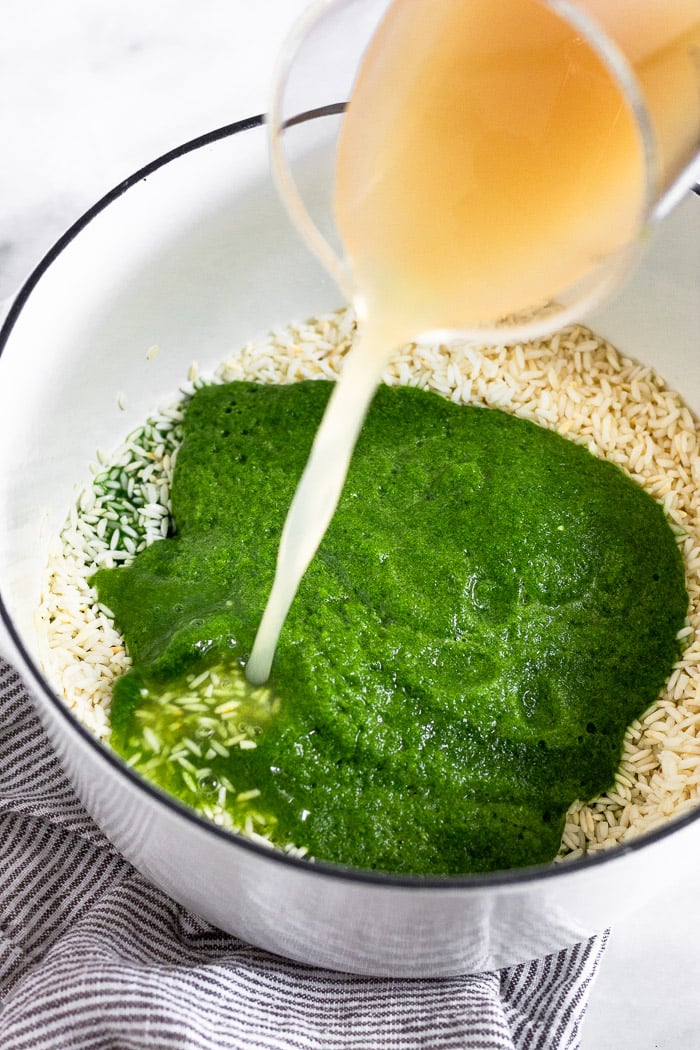 Large pot with toasted rice, green broth mixture, and broth being poured into it to make a green rice recipe.