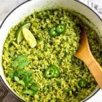 Large dutch oven filled with arroz verde topped with limes, jalapenos, and cilantro. There is a wooden spoon coming out of the pan. The pan is sitting on a towel with a bowl of cilantro and limes next to it.