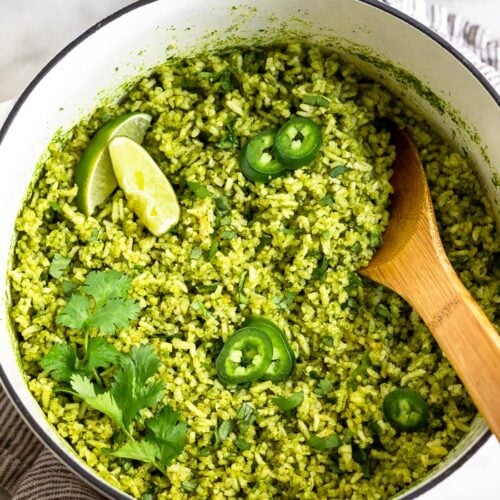 Large dutch oven filled with arroz verde topped with limes, jalapenos, and cilantro. There is a wooden spoon coming out of the pan. The pan is sitting on a towel with a bowl of cilantro and limes next to it.