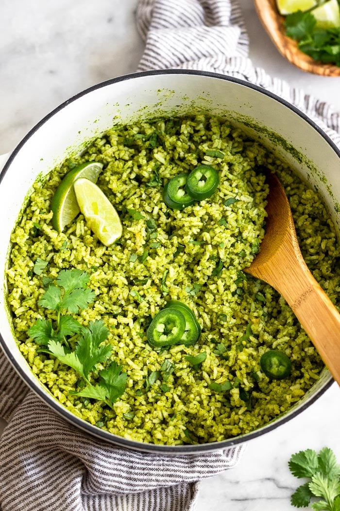 Large dutch oven filled with arroz verde topped with limes, jalapenos, and cilantro. There is a wooden spoon coming out of the pan. The pan is sitting on a towel with a bowl of cilantro and limes next to it.