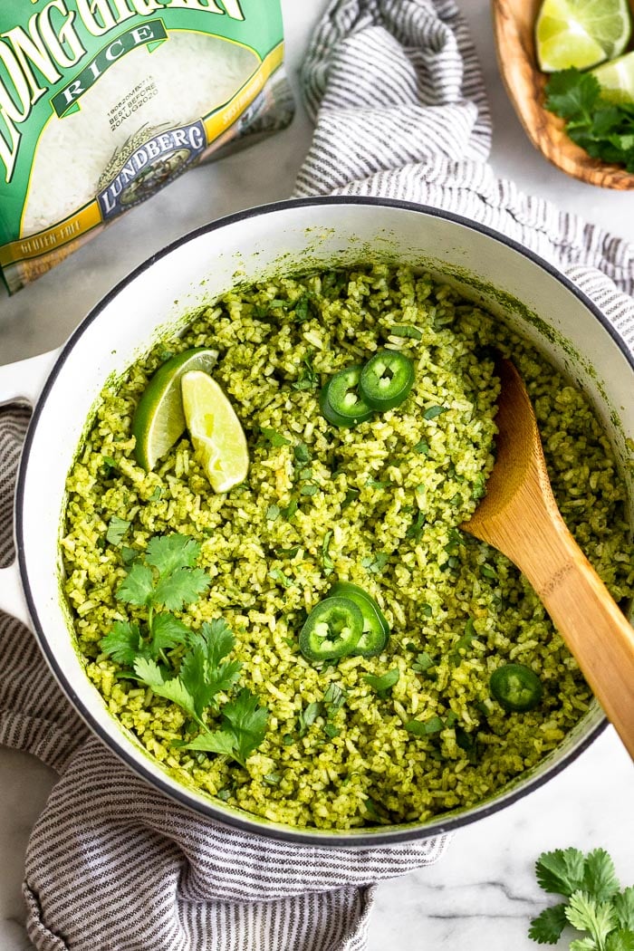 Large stock pot filled with green rice topped with jalapeños, limes wedges, and cilantro. Around it is a bag of white rice, a plate of herbs and lime, and a striped linen.