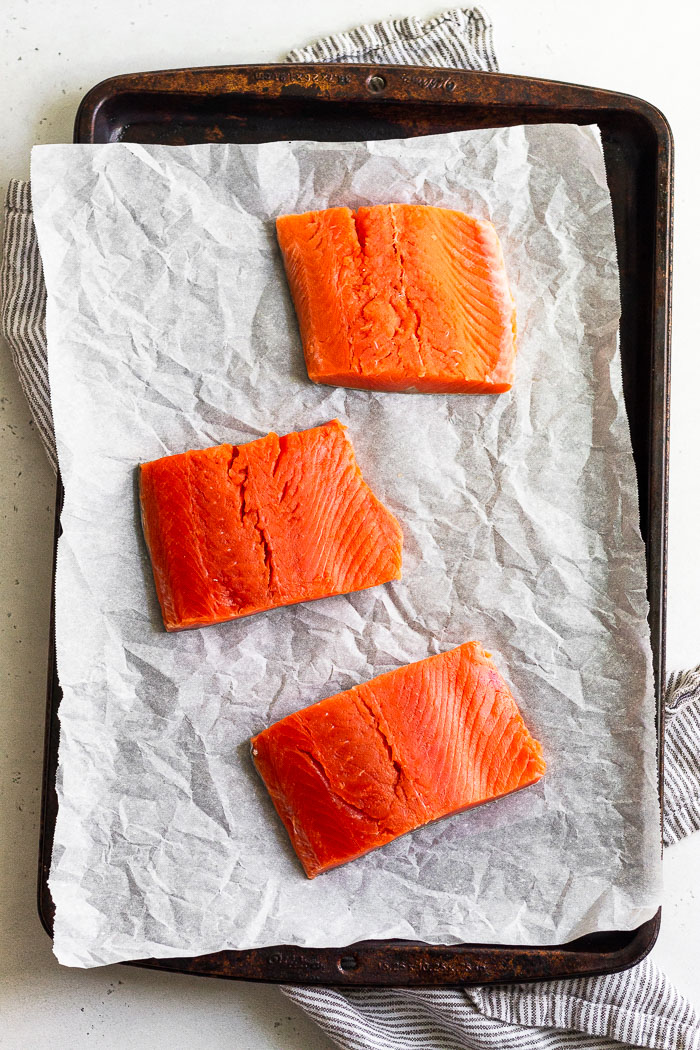Overhead view of 3 raw salmon filets on a baking sheet lined with parchment paper.
