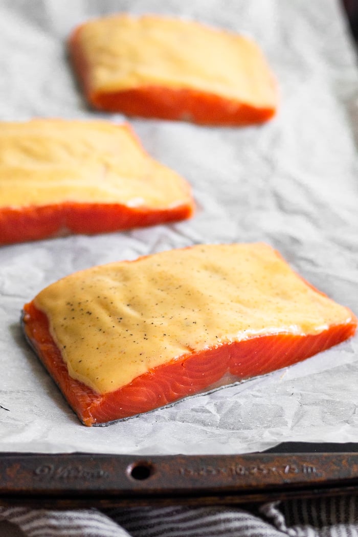 Baking sheet lined with parchment paper with 3 raw salmon filets topped with chipotle mayo.