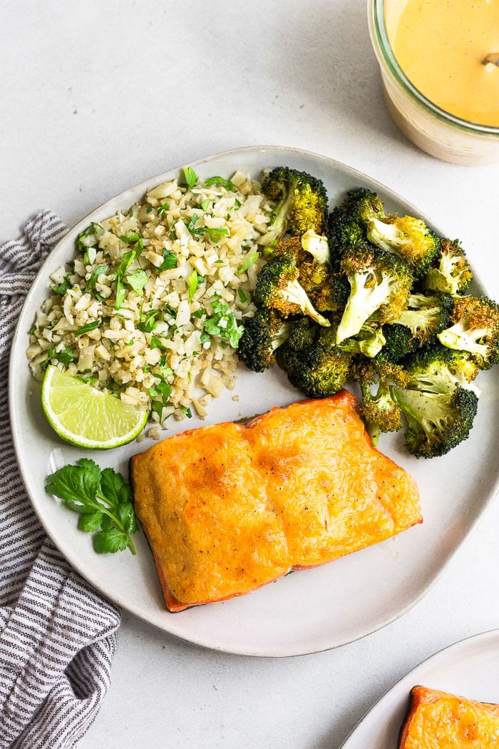 Plate of keto salmon next to roasted broccoli and cilantro lime cauliflower rice. Next to it is a jar of homemade chipotle mayo and another plate, both of them being halfway out of the picture.
