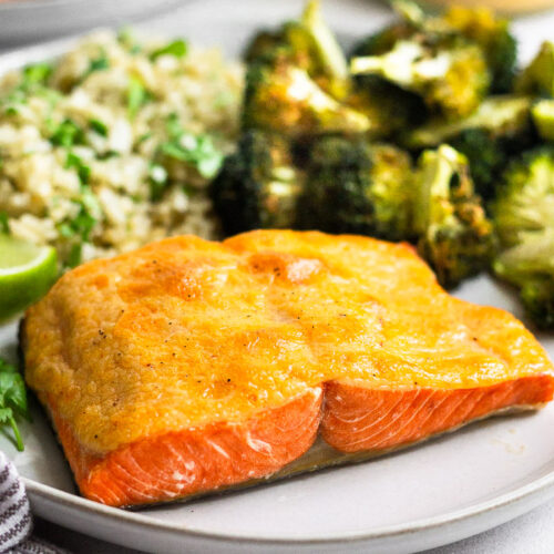 Plate of air fryer salmon that is covered in chipotle mayo. Next to it is roasted broccoli and cauliflower rice. Behind it is the jar of mayo and another plate of salmon.
