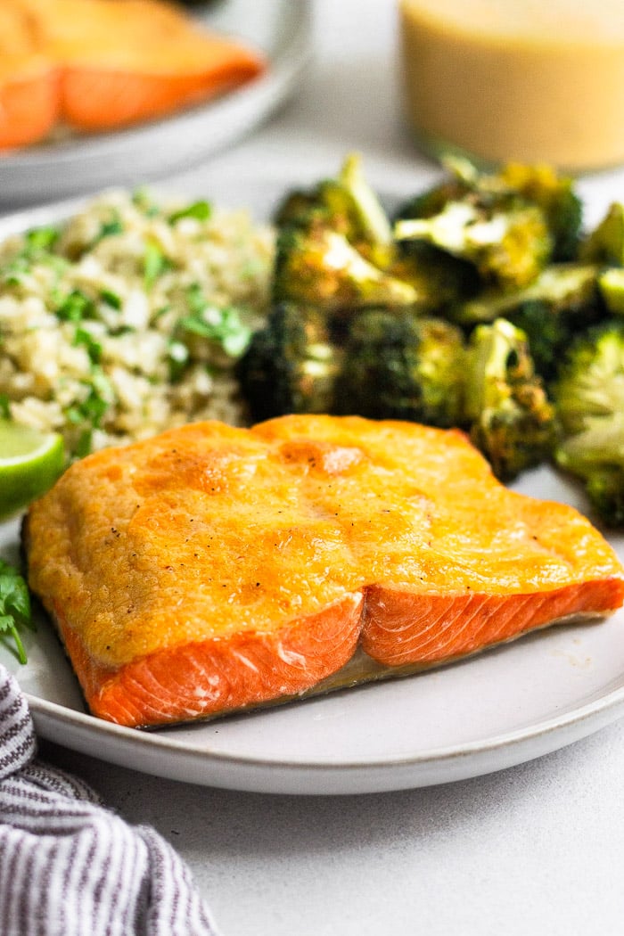 Plate of air fryer salmon that is covered in chipotle mayo. Next to it is roasted broccoli and cauliflower rice. Behind it is the jar of mayo and another plate of salmon.