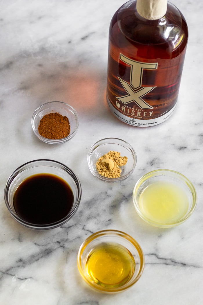 White counter top with a bottle of whiskey, and small bowls filled with coconut aminos, lemon juice, honey, cinnamon, and ground ginger
