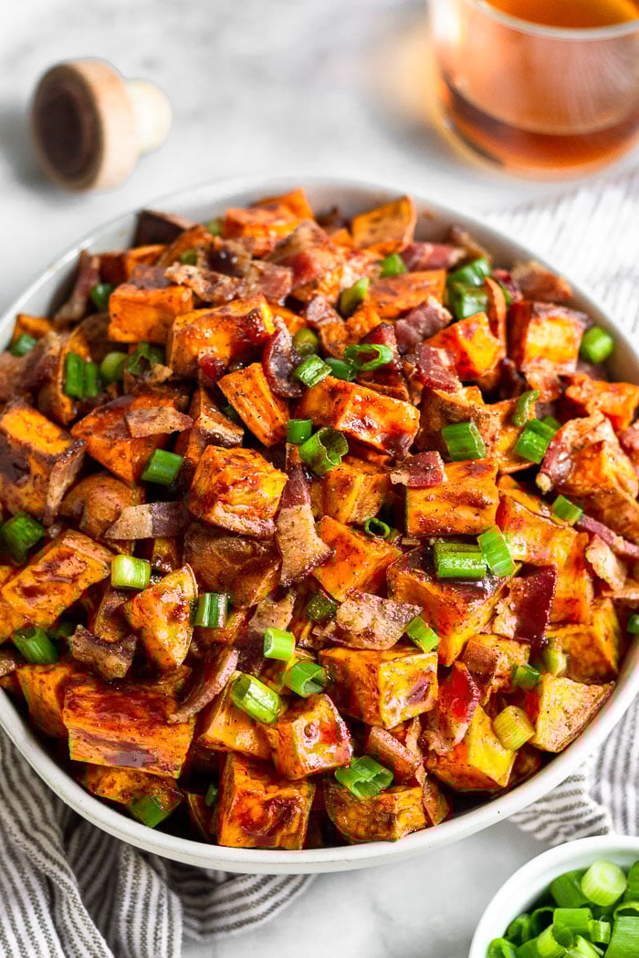 A bowl of glazed sweet potatoes with a glass of whiskey, a bowl of green onion, and a cork to a bottle around it.