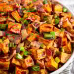 Bowl of glazed sweet potatoes topped with bacon and green onions. Next to is a bowl of green onions and behind it a glass of whiskey.